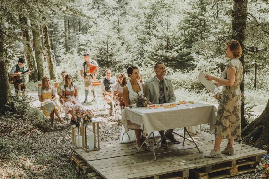 Trauung an der Waldlichtung. Hochzeitspaar und Gäste sitzen vor der Standesbeamtin.