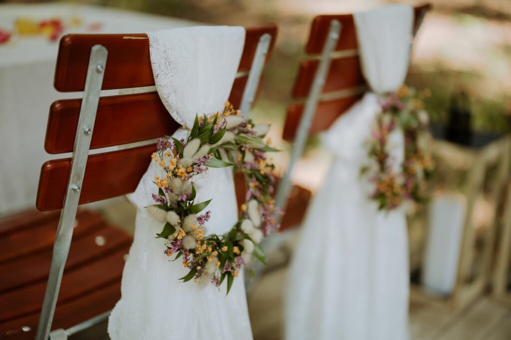 Zwei Stühle mit weißen Tüchern und bunten Blumenkränzen dekoriert.